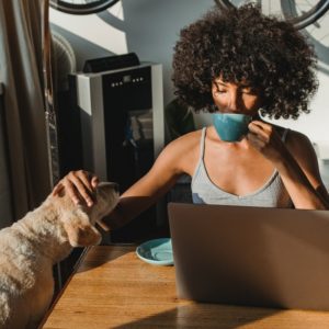 A black woman at her desk, behind her laptop. She is drinking from a cup and patting her dog.